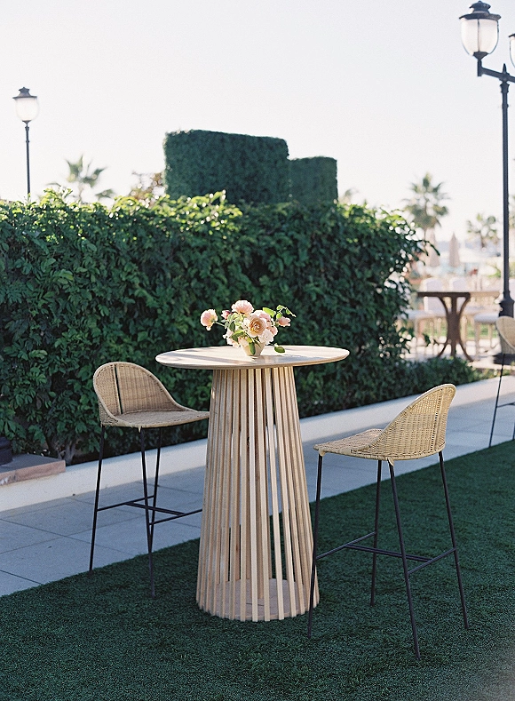 Cocktail table setup with outdoor cocktail table decor, small floral centerpiece on a round high-top beside rattan stools on a hedge-lined terrace
