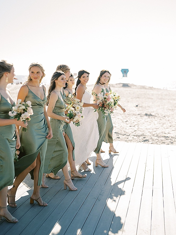 Bridesmaids portrait of bride with bridesmaids walking in sage green satin dresses holding bouquets on a beach by the ocean horizon