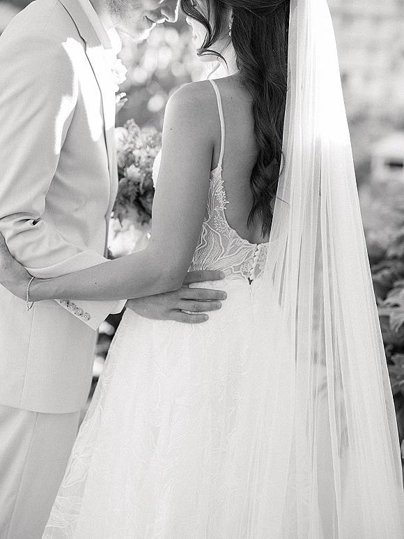 Wedding couple portrait of bride and groom embrace, nose-to-nose, her long veil and lace dress beside his white suit in greenery