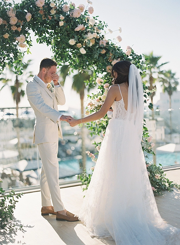 First look moment as bride in a veil and wedding dress holds groom’s hand on a marina balcony with palm trees and boats behind