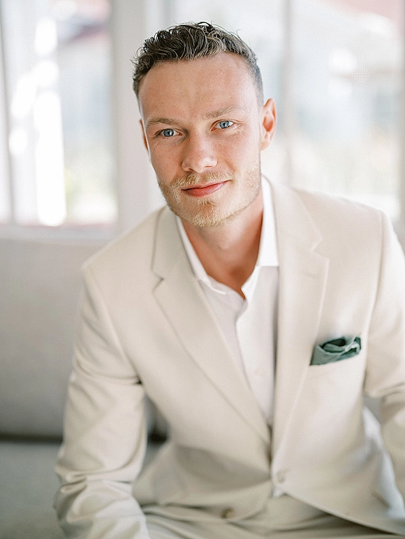 Groom portrait in soft window light, beige suit with white open-collar shirt and pocket square, standing indoors by blurred windows
