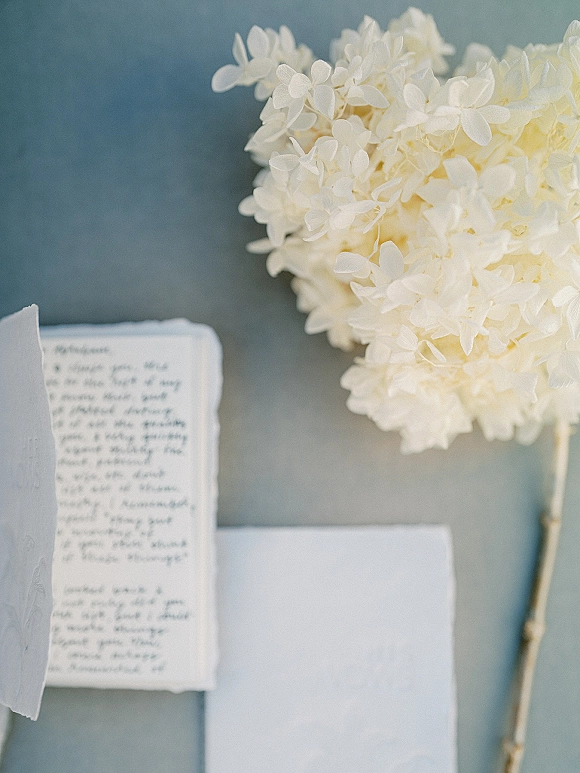 Wedding invitation suite with deckle edge wedding invitations, white hydrangea accent, envelope, and handwritten note on a gray tabletop
