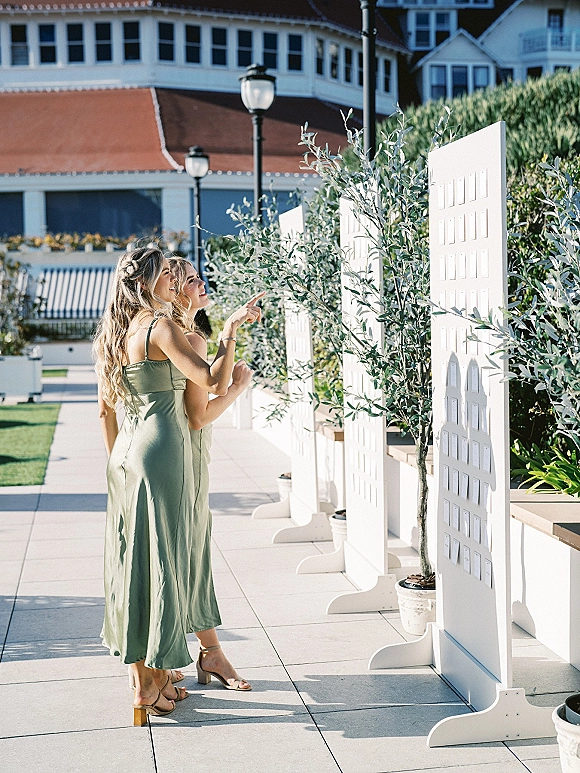 Wedding seating chart with escort card display on white boards as bridesmaids in sage dresses browse on a hotel patio with potted olive trees