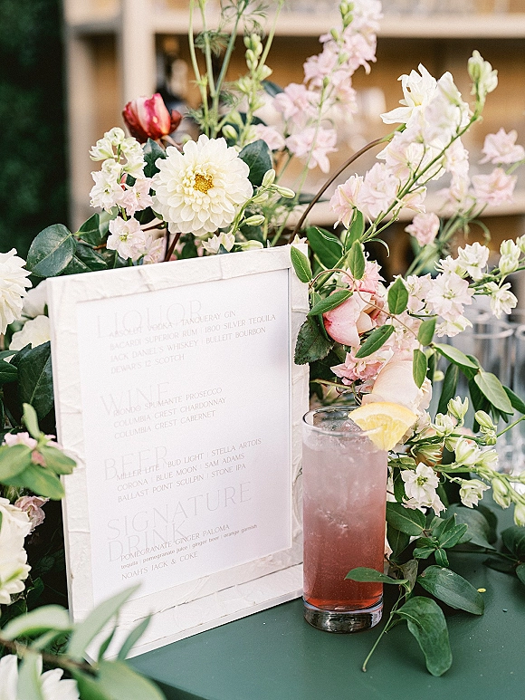 Wedding bar menu and signature drink sign in a white frame with pink roses and greenery, set on a bar counter with shelves of bottles behind