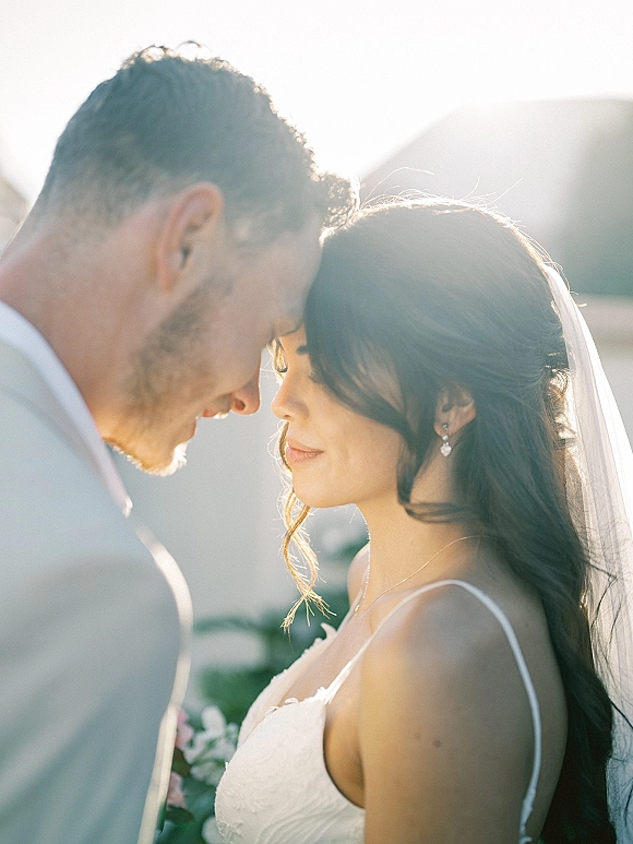 Couple portrait of bride and groom close up touching foreheads, her veil and bouquet backlit at golden hour with greenery and sky behind