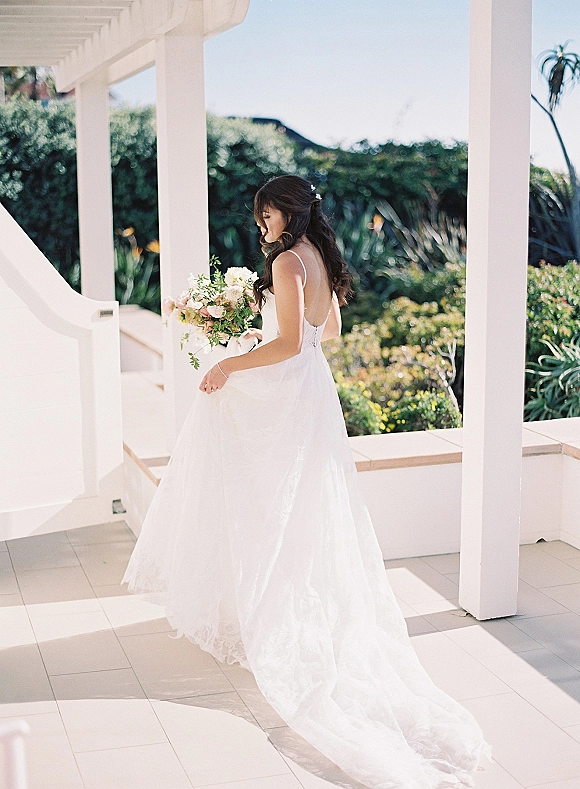 Bridal portrait of bride holding bouquet, showing a backless wedding dress with lace hem on a white porch with columns and greenery