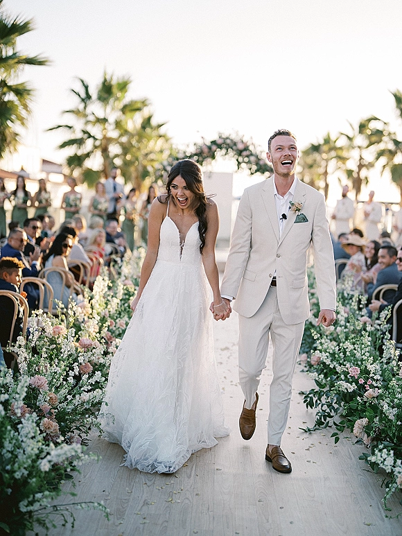 Wedding recessional as bride and groom walking aisle holding hands, laughing past cheering guests on a palm tree outdoor ceremony path