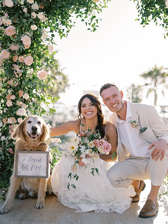 Couple portrait of bride and groom with dog, bride holding bouquet beside a floral arch on a wooden deck with palm trees behind
