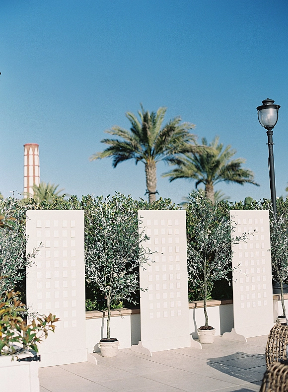 Wedding escort display with white escort card panels and potted trees against a greenery hedge on an outdoor patio under palm trees and blue sky