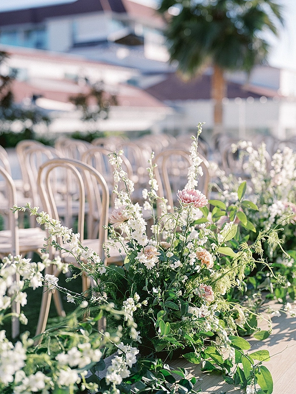 Ceremony aisle florals with white and blush blooms and greenery lining wooden chairs on an outdoor lawn beneath palm trees and sky