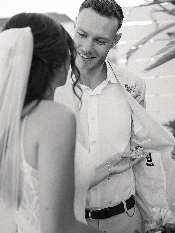 Wedding couple portrait with groom smiling at bride as she adjusts his jacket, bridal veil and boutonniere under indoor lights and plants