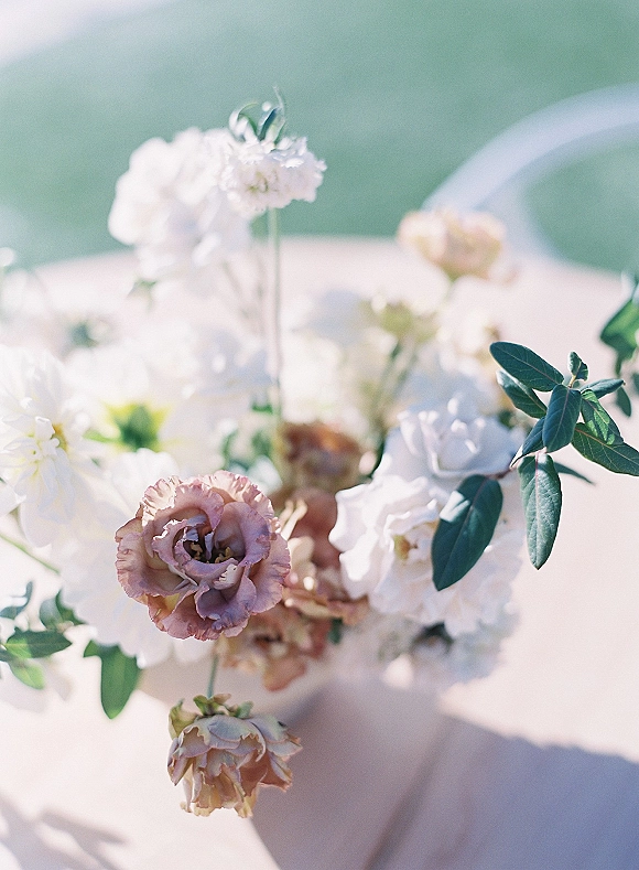Wedding centerpiece with a low floral centerpiece of white and blush flowers with greenery on a tablecloth, outdoor greenery behind