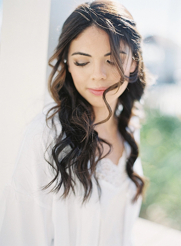 Bridal portrait of a bride looking down in soft window light, wearing a lace bodice wedding dress and veil, with blurred greenery behind