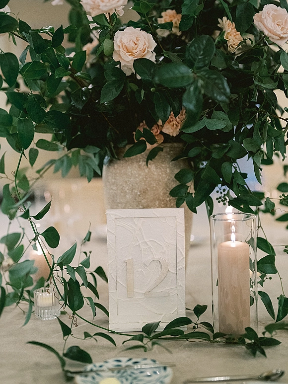 Reception centerpiece with blush roses and greenery in a textured vase, flanked by pillar and votive candles beside a table number card