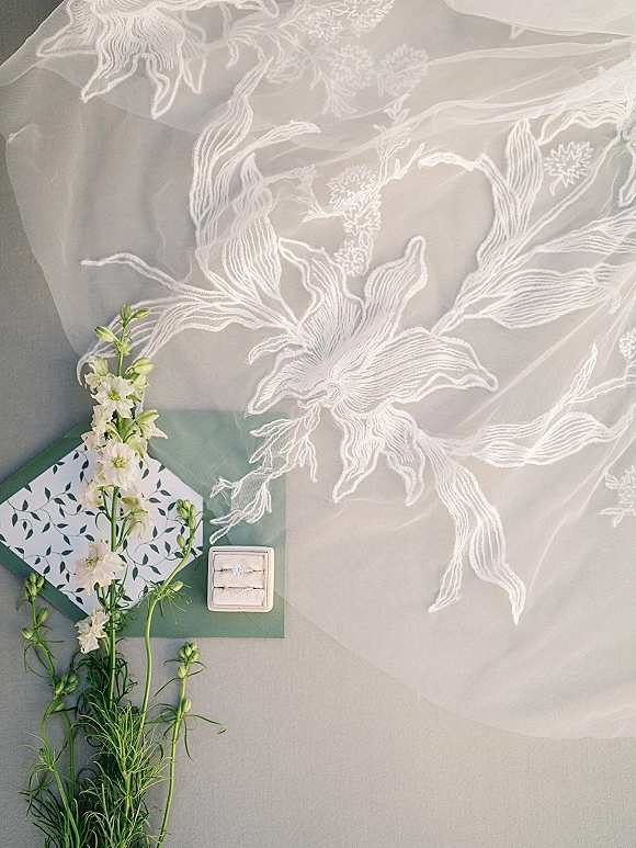Wedding rings flatlay featuring an engagement ring and band in a square ring box with lace veil and stationery on a gray surface