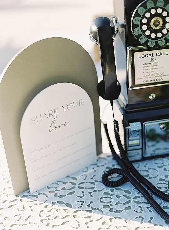 Wedding audio guestbook with audio guestbook phone, a vintage payphone on lace tablecloth beside a clear arch sign in soft outdoor light