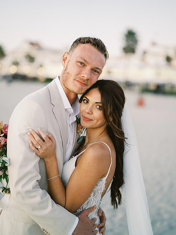 Couple portrait of bride and groom embrace, her veil and lace dress visible as he holds her by the ocean shoreline under open sky