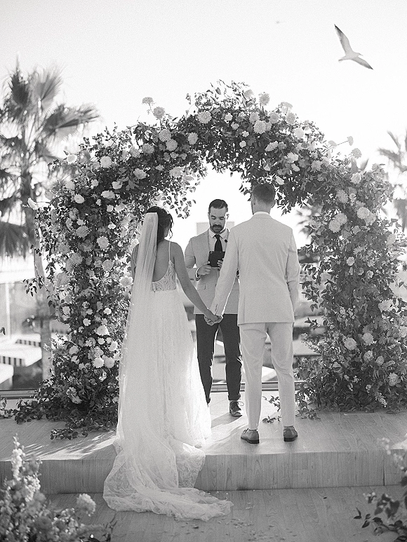 Wedding ceremony moment as bride and groom hold hands under a round floral arch on a terrace with palm trees and ocean view backdrop