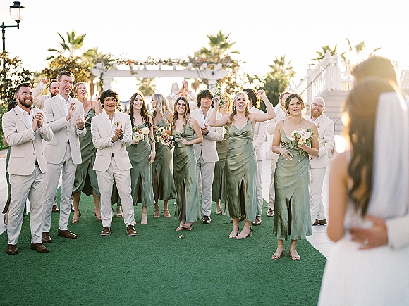Wedding party photo of bridesmaids and groomsmen photo cheering in sage green dresses and light beige suits under a greenery pergola with palm trees behind