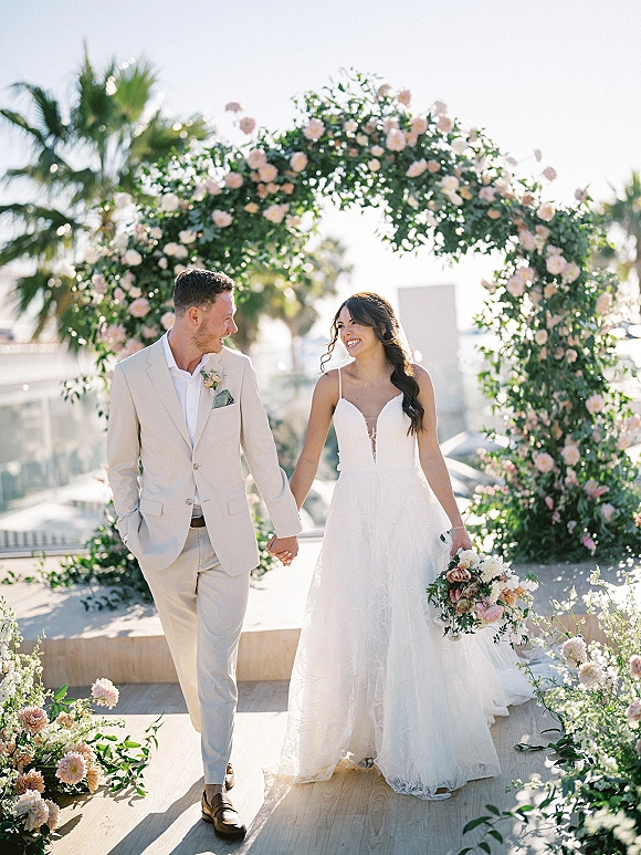 Couple portrait of bride in deep V wedding dress and groom in light tan suit holding hands beneath a rose floral arch on an oceanfront terrace