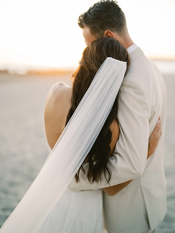 Couple portrait of the bride and groom embrace as a wedding veil blows in the breeze, facing the ocean horizon on a beach