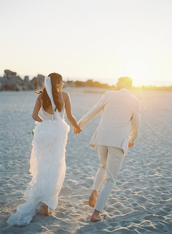 Couple portrait of bride and groom holding hands walking away barefoot on a sandy beach at sunset, veil blowing by the ocean horizon