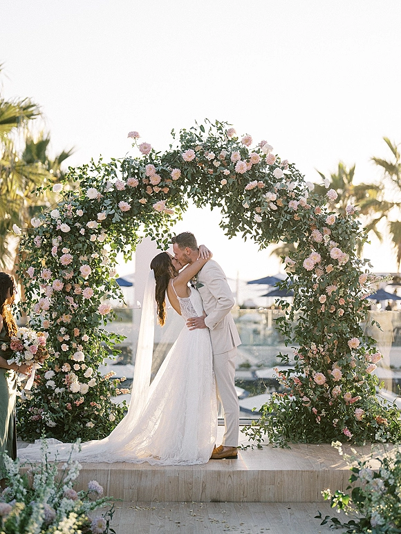 Wedding kiss at first kiss ceremony under a round floral arch with pink flowers, bride’s veil and groom in light suit, palm trees behind