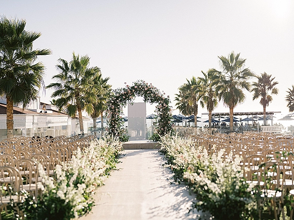 Ceremony setup with a round floral arch and white aisle flowers, wooden chairs facing the ocean on a palm-lined beach club patio