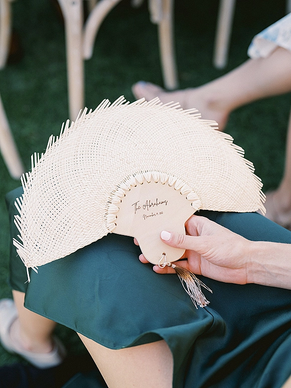 Wedding ceremony fan held by a guest in a dark green dress, a custom wedding fan with tassel and calligraphy on a grassy lawn with white chairs