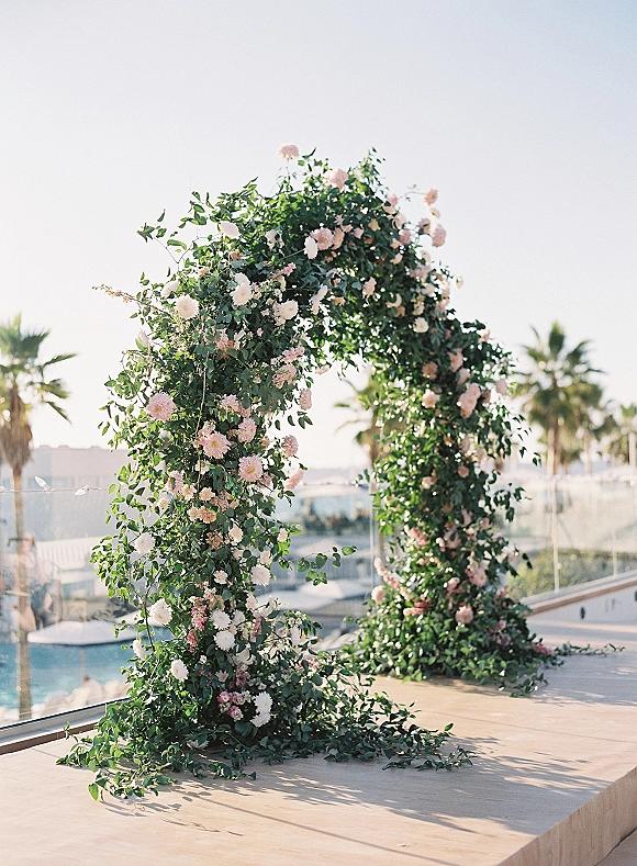 Floral wedding arch with lush greenery garland and blush and white blooms on an outdoor terrace, framed by glass railing and palm trees