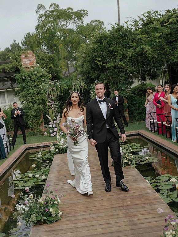Recessional moment as bride and groom walking aisle hand in hand, bouquet and tuxedo, over a wooden walkway by a lily pond under overcast skies