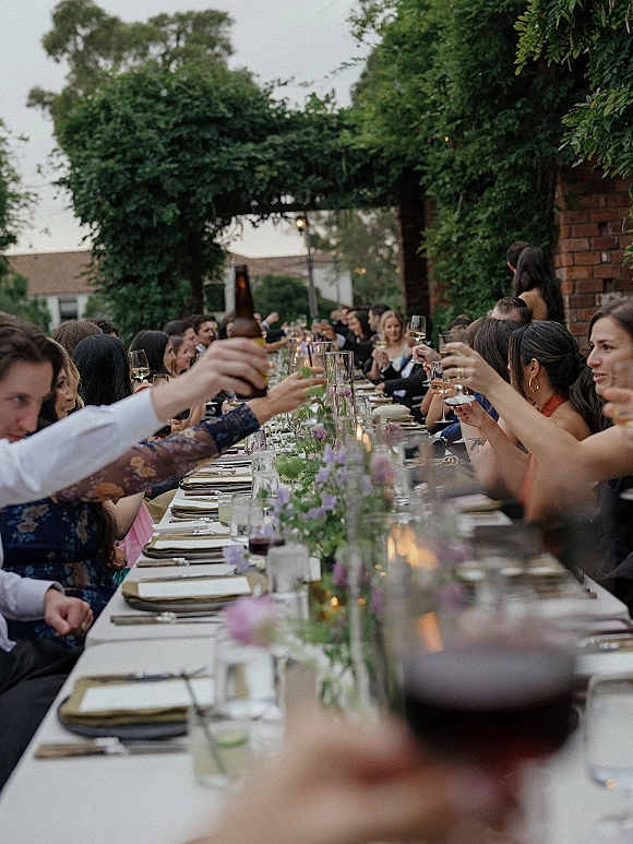 Wedding reception toast as guests raise wine glasses and a beer bottle over a candlelit banquet table under string lights in a brick courtyard