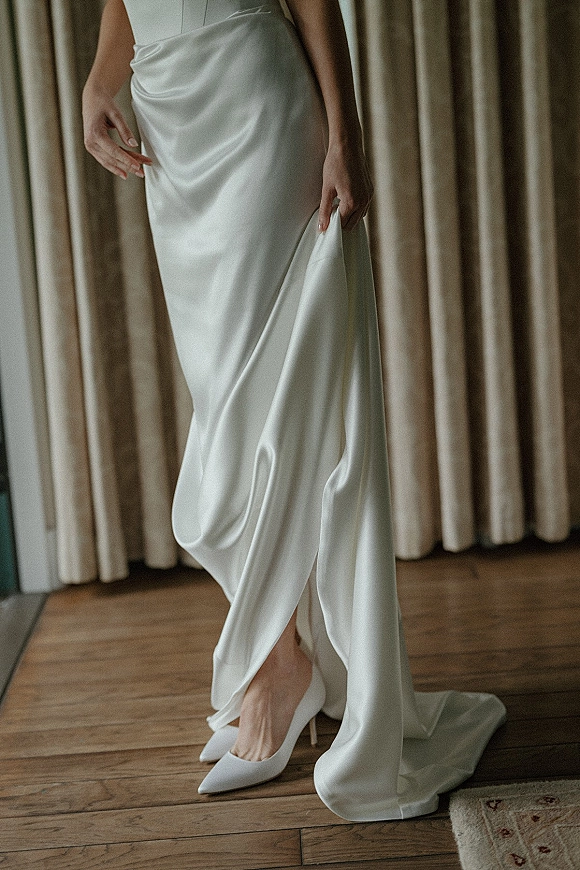 Wedding dress close-up with satin wedding dress draping over a skirt beside pointed toe bridal heels on a rug over wooden floor