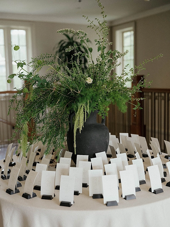 Escort card display with wedding escort cards in black holders beside a black ceramic vase of greenery and white flowers in an indoor room