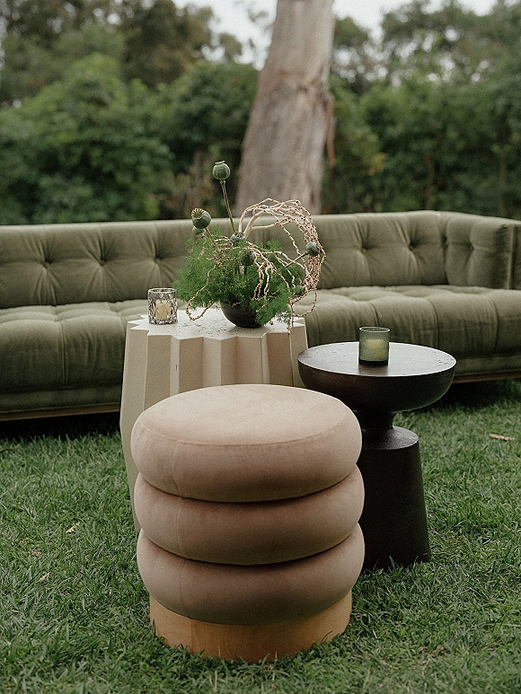 Lounge seating with a velvet tufted sofa, round side tables, candles, and floral centerpiece on a garden lawn with trees