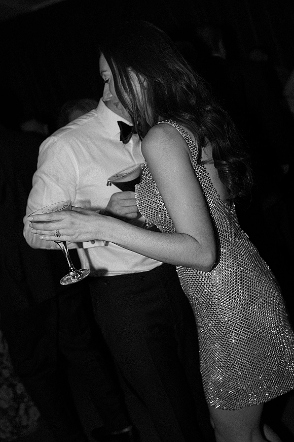 Wedding reception moment with wedding party dancing, showing a couple in sequin mini dress and bow tie holding a cocktail in a dark crowd