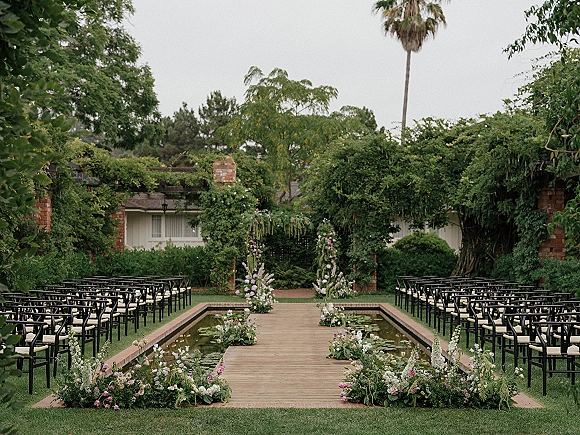 Ceremony aisle design with a wood plank walkway over a reflecting pool, lined with pink and white florals and garden chairs under a pergola