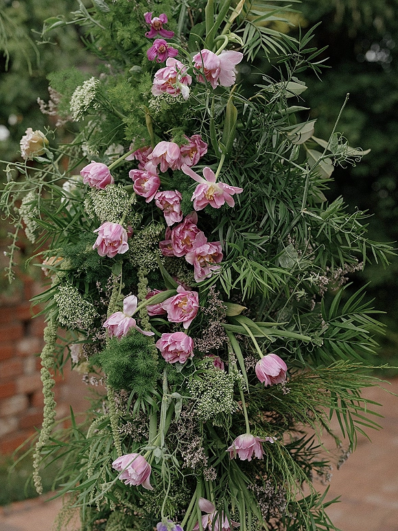Wedding floral installation with pink tulip accents and trailing greenery, cascading asymmetrically against garden foliage and a brick wall