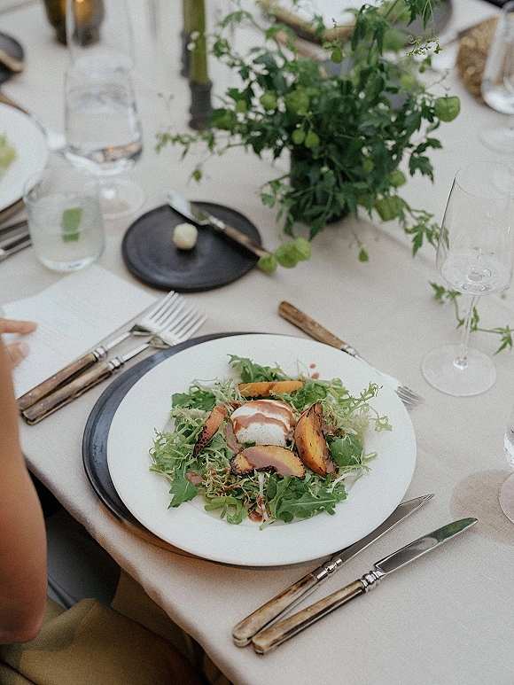 Reception tablescape with wedding place setting, white plate on black charger, salad course, flatware, glasses, greenery and taper candles on linen tablecloth