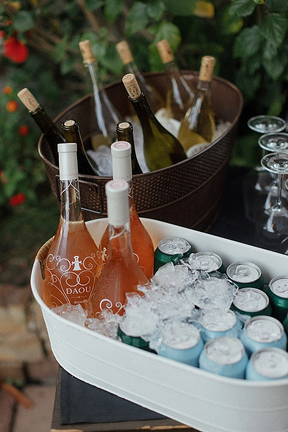 Wedding drink station with wine bottles and rosé on ice, beer cans, and coupe glasses on a table surrounded by greenery