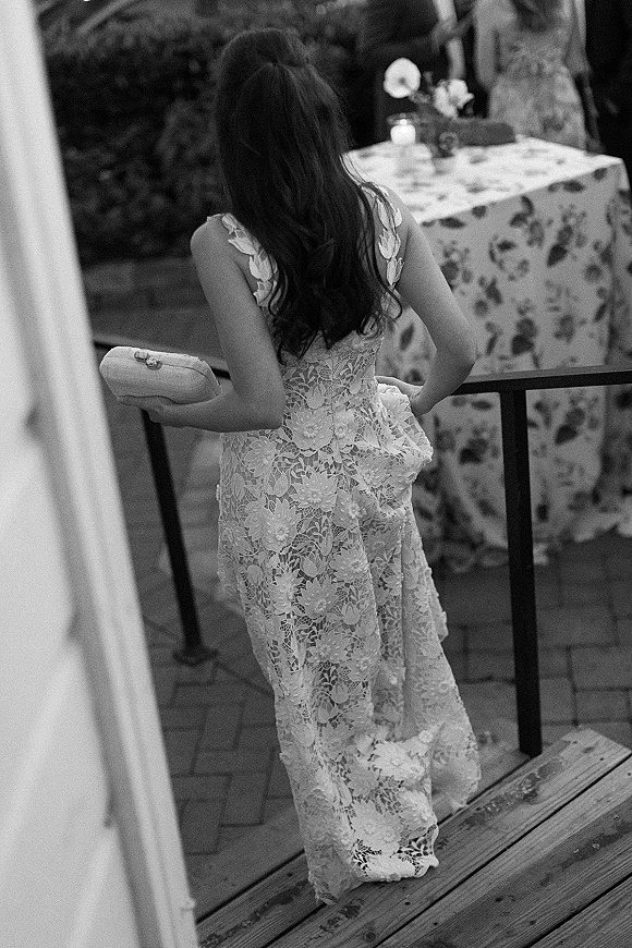 Wedding guest candid in a long lace maxi dress holding a clutch by a cocktail table on a brick patio with wooden steps and greenery