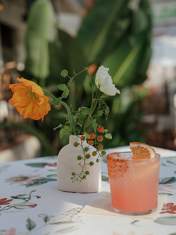 Reception tablescape with a pink grapefruit cocktail beside a bud vase of orange and white blooms on a printed tablecloth outdoors