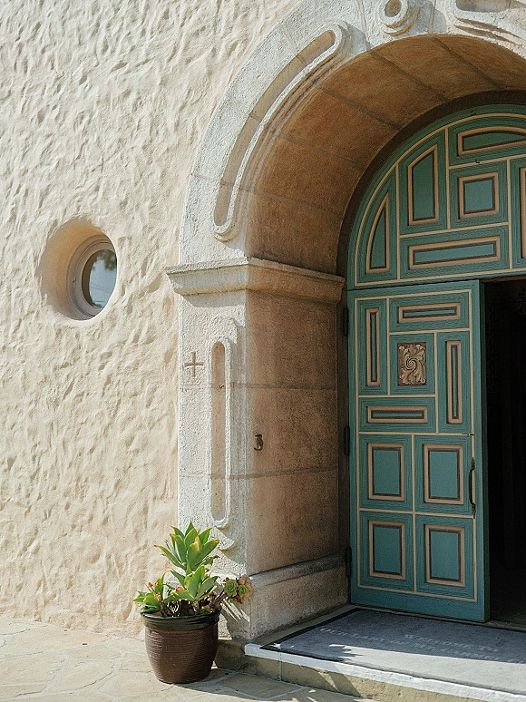 Chapel entrance with a potted succulent accent and doorway rug beneath a stone archway, set against a stucco wall and open doorway