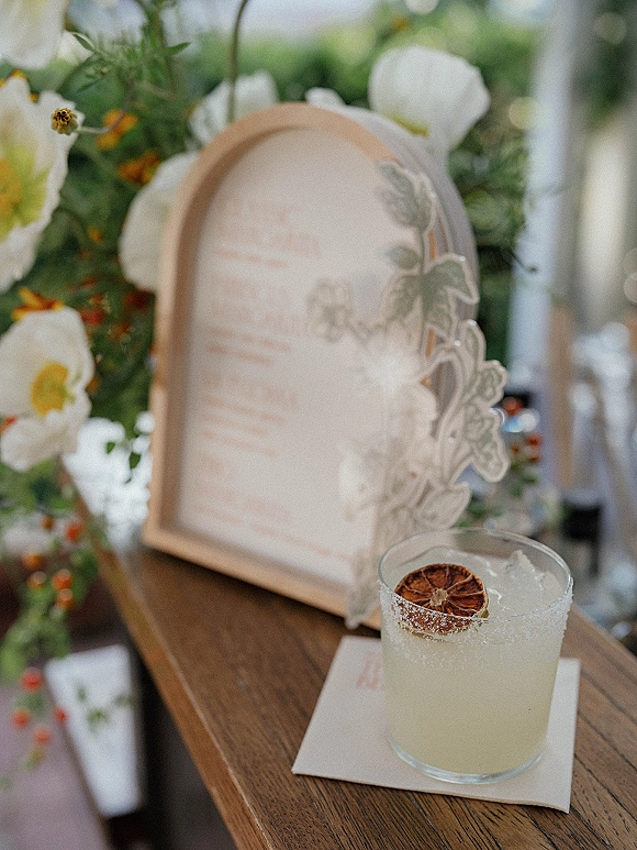 Signature wedding cocktail with a salt rim and dried citrus garnish beside a signature drink sign on a wood bar top with greenery behind