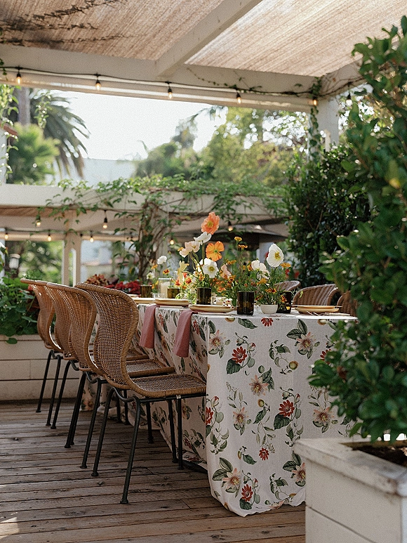 Reception tablescape with floral tablecloth, wildflower centerpieces, taper candles and glass votives on an outdoor patio under string lights