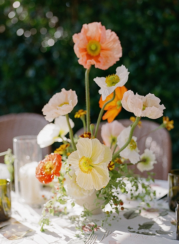 Wedding centerpiece with poppy wedding centerpiece blooms, greenery, candles and menu card on patterned tablecloth with bistro chairs behind