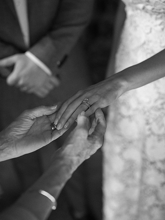 Wedding ring exchange close up as groom places wedding band on bride’s hand, diamond engagement ring and manicure visible against blurred attire