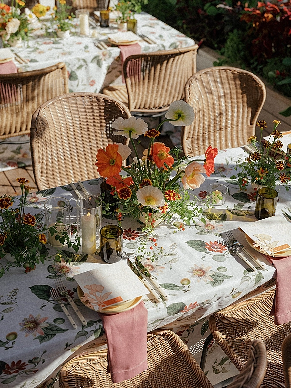 Reception tablescape with a floral print tablecloth, wildflower centerpieces, taper candles, pink napkins, and wicker chairs on a leafy patio