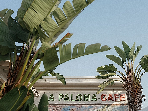 Wedding venue exterior with neon sign glowing above a painted mural, framed by palm trees against a bright blue sky