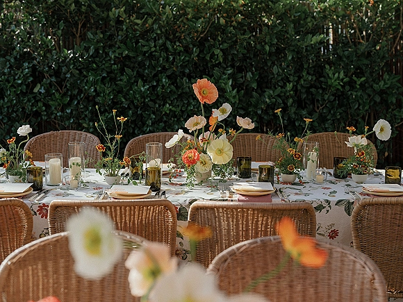 Reception tablescape with an outdoor reception table of wildflower centerpieces, amber glass tumblers, and pillar candles set before a green hedge
